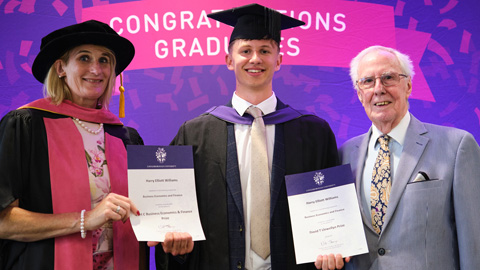 Three people at a graduation ceremony; two in academic regalia holding certificates, one in formal attire.